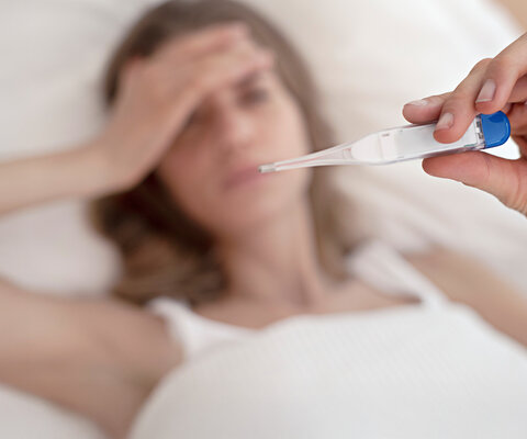 Eine liegende Frau hält ein Thermometer mit der Hand in die Luft, während die andere Hand auf ihrer Stirn liegt.