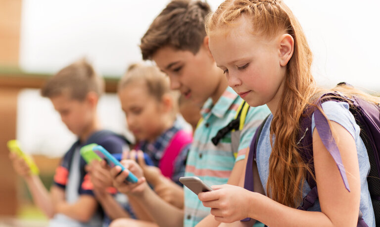 Vier Schulkinder mit Rucksäcken, die alle ein Smartphone in der Hand halten, auf welches sie blicken.