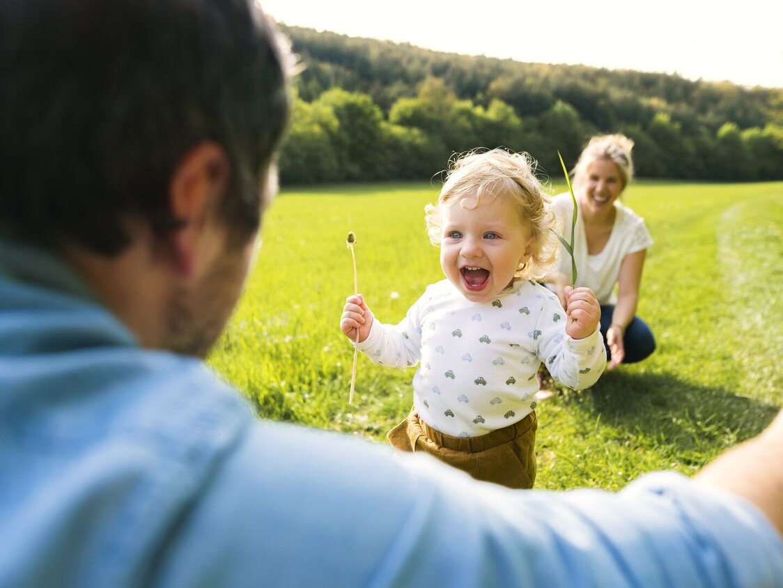 Ein Kleinkind läuft auf einer WIese aus Richtung einer Frau in die Arme eines Mannes.