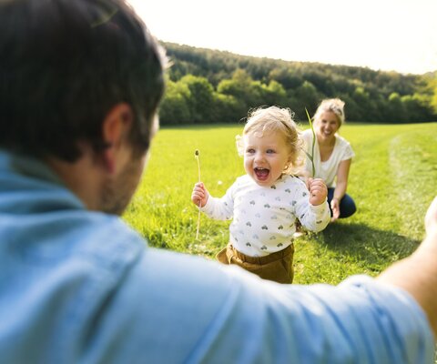 Ein Kleinkind läuft auf einer WIese aus Richtung einer Frau in die Arme eines Mannes.