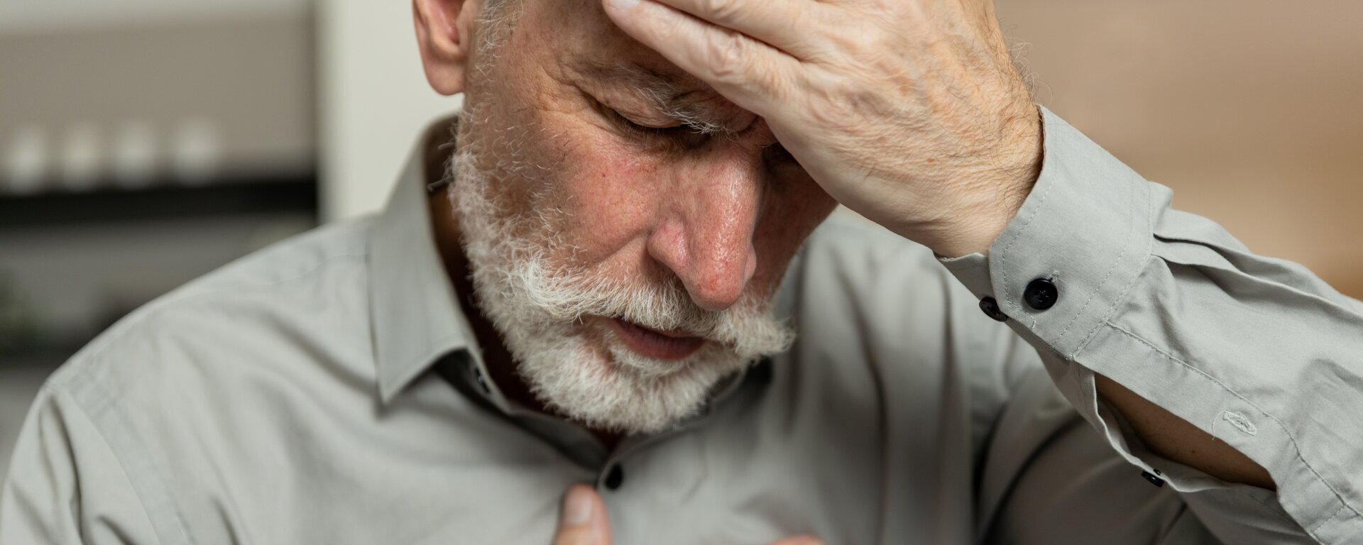 Ein älterer Mann hält sich mit einer Hand die Stirn, drückt mit der anderen auf die Brust, sieht verzweifelt aus und zeigt Anzeichen von Schmerzen und Erschöpfung. Schwindel, gesundheitliche Probleme. Psychische Störung, Herzerkrankung
