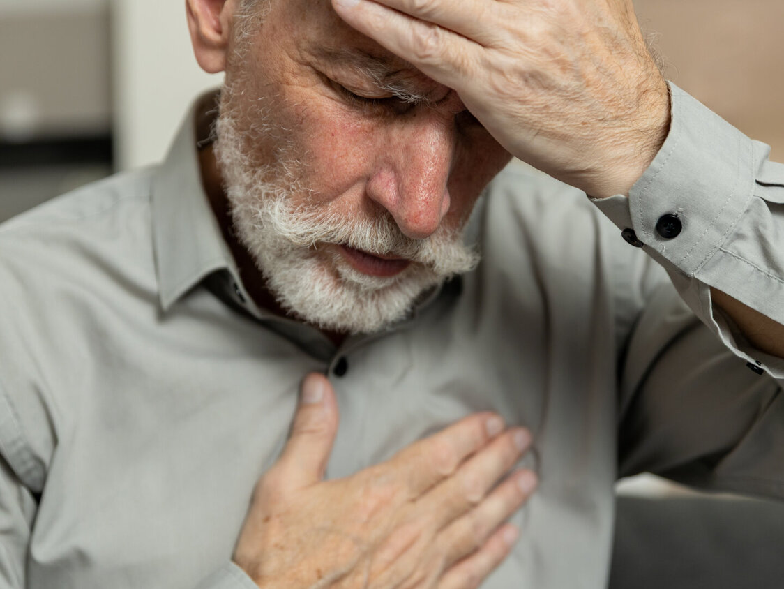 Ein älterer Mann hält sich mit einer Hand die Stirn, drückt mit der anderen auf die Brust, sieht verzweifelt aus und zeigt Anzeichen von Schmerzen und Erschöpfung. Schwindel, gesundheitliche Probleme. Psychische Störung, Herzerkrankung
