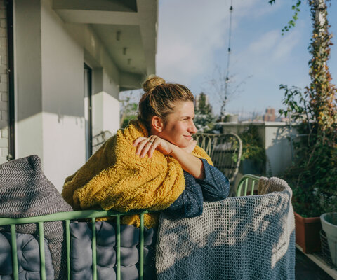 Eine Frau lehnt gegen ein Balkongeländer und hält ihr Gesicht in die Sonne.