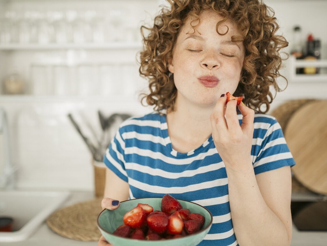 Eine Frau mit roten Locken steht  in der Küche und ist Erdbeeren aus einer Schale. Dabei grinst sie..