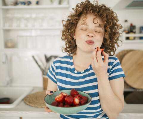 Eine Frau mit roten Locken steht  in der Küche und ist Erdbeeren aus einer Schale. Dabei grinst sie..