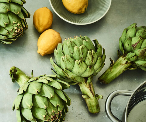 Auf einem Edelstahluntergrund liegen sechs Artischocken, dazwischen drei Zitronen und am Rand steht ein Kochtopf.