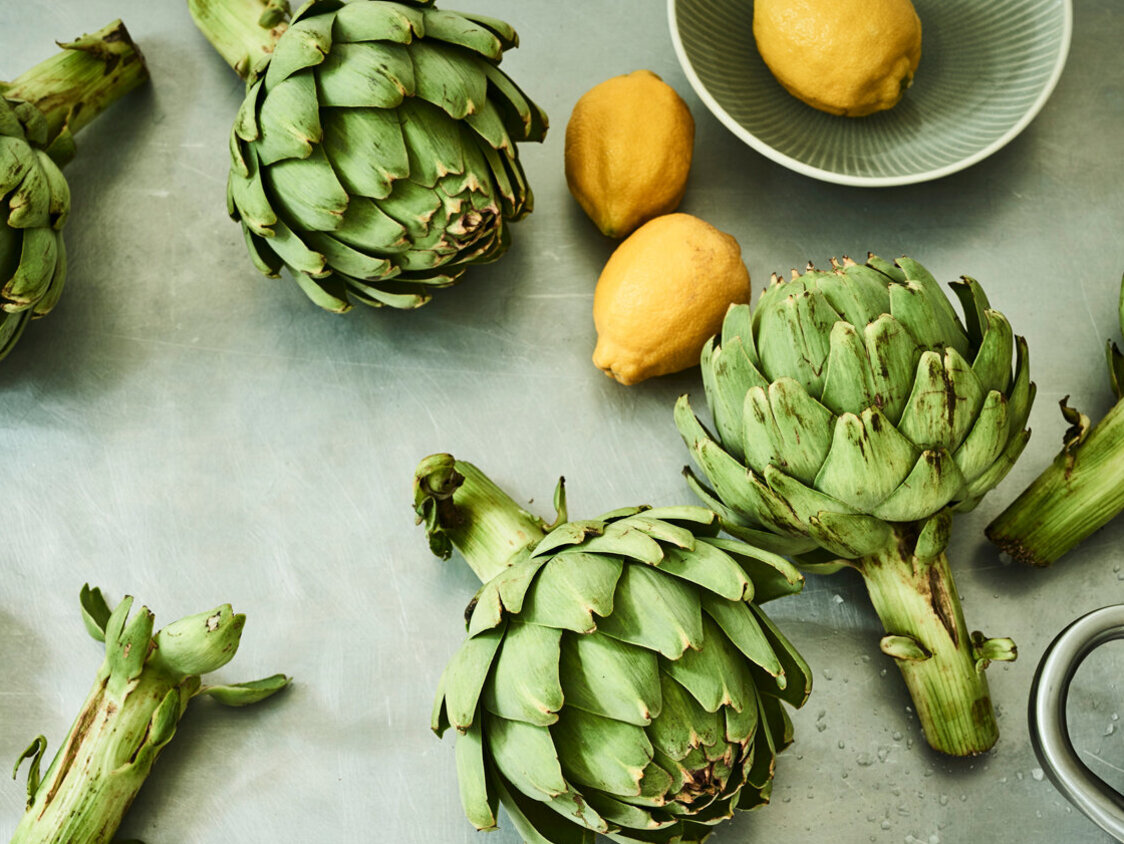 Auf einem Edelstahluntergrund liegen sechs Artischocken, dazwischen drei Zitronen und am Rand steht ein Kochtopf.