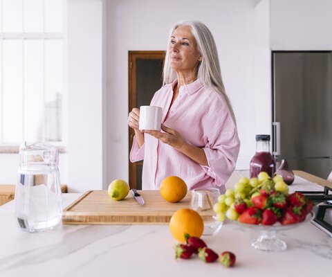 Eine ältere Frau steht in der Küche vor einem Schneidebrett mit Zitrusfrüchten und hält eine Tasse in der Hand. 
