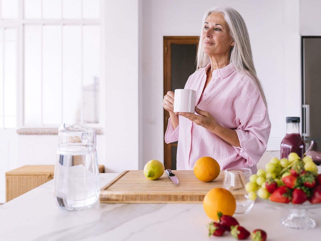 Eine ältere Frau steht in der Küche vor einem Schneidebrett mit Zitrusfrüchten und hält eine Tasse in der Hand. 