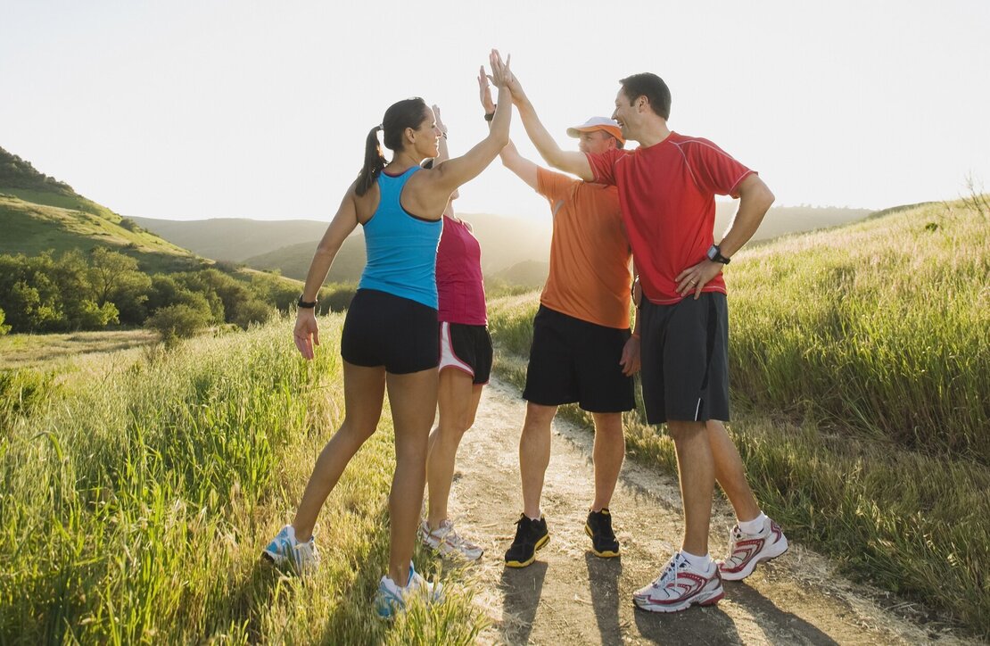 Laufgruppe von vier Personen in Sportbekleidung klatscht sich auf einem Weg durch ein grünes Feld ab.