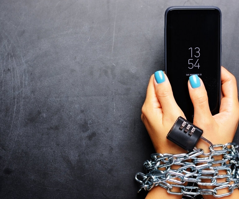 Zwei Hände die ein Smartphone halten und mit Stahlketten und einem Schloss gefesselt sind.