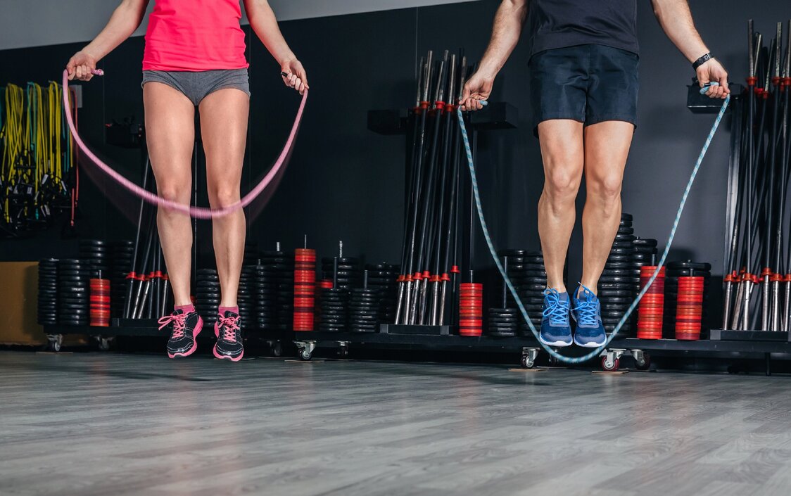  Ein Mann und eine Frau beim Seilspringen. Man sieht nur ihre Beine. Sie sind in einem Fitnessraum, hinter ihnen stehen Fitnessgeräte.