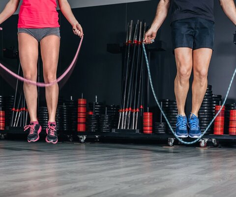  Ein Mann und eine Frau beim Seilspringen. Man sieht nur ihre Beine. Sie sind in einem Fitnessraum, hinter ihnen stehen Fitnessgeräte.