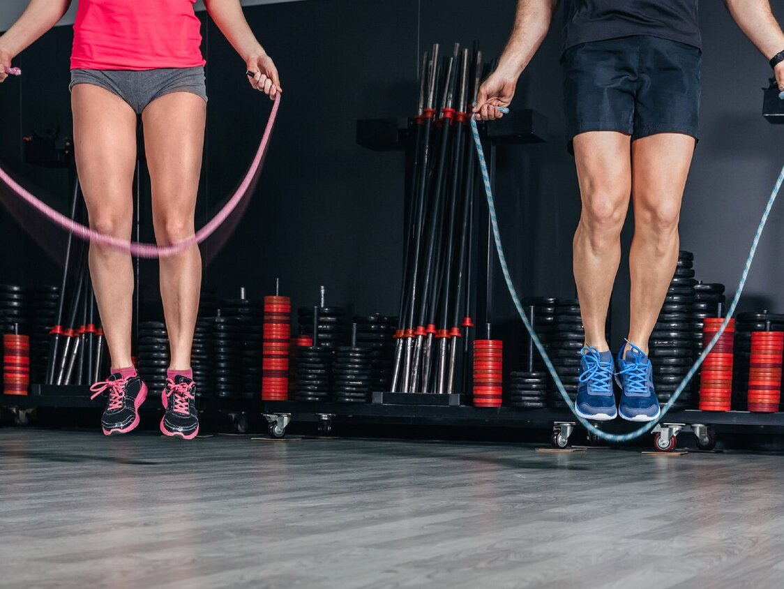 Ein Mann und eine Frau beim Seilspringen. Man sieht nur ihre Beine. Sie sind in einem Fitnessraum, hinter ihnen stehen Fitnessgeräte.