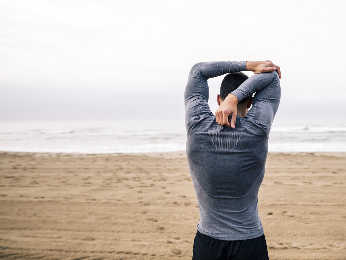 Ein Mann steht am Strand und dehnt seinen Arm.