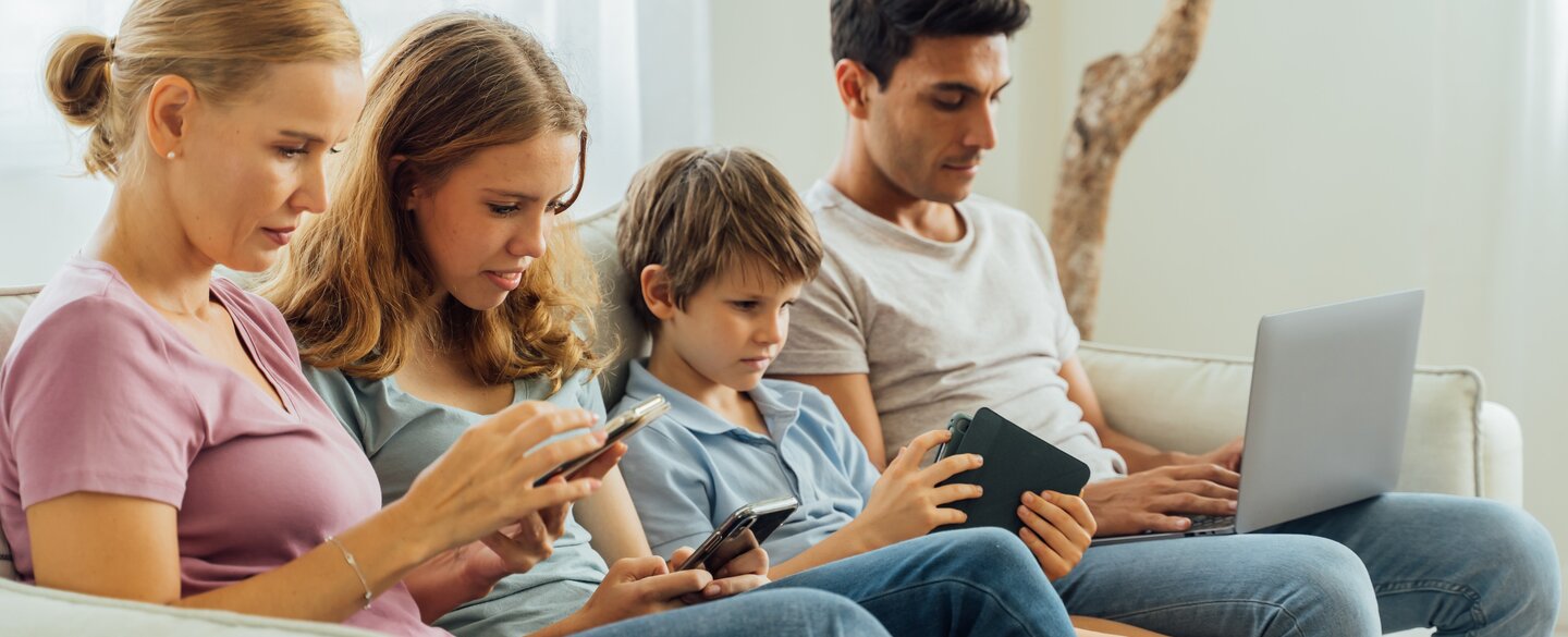 Eine Familie, welche auf einem Sofa sitzt. Jedes Familienmitglied hält ein mobiles Endgerät in den Händen, auf welches es blickt.