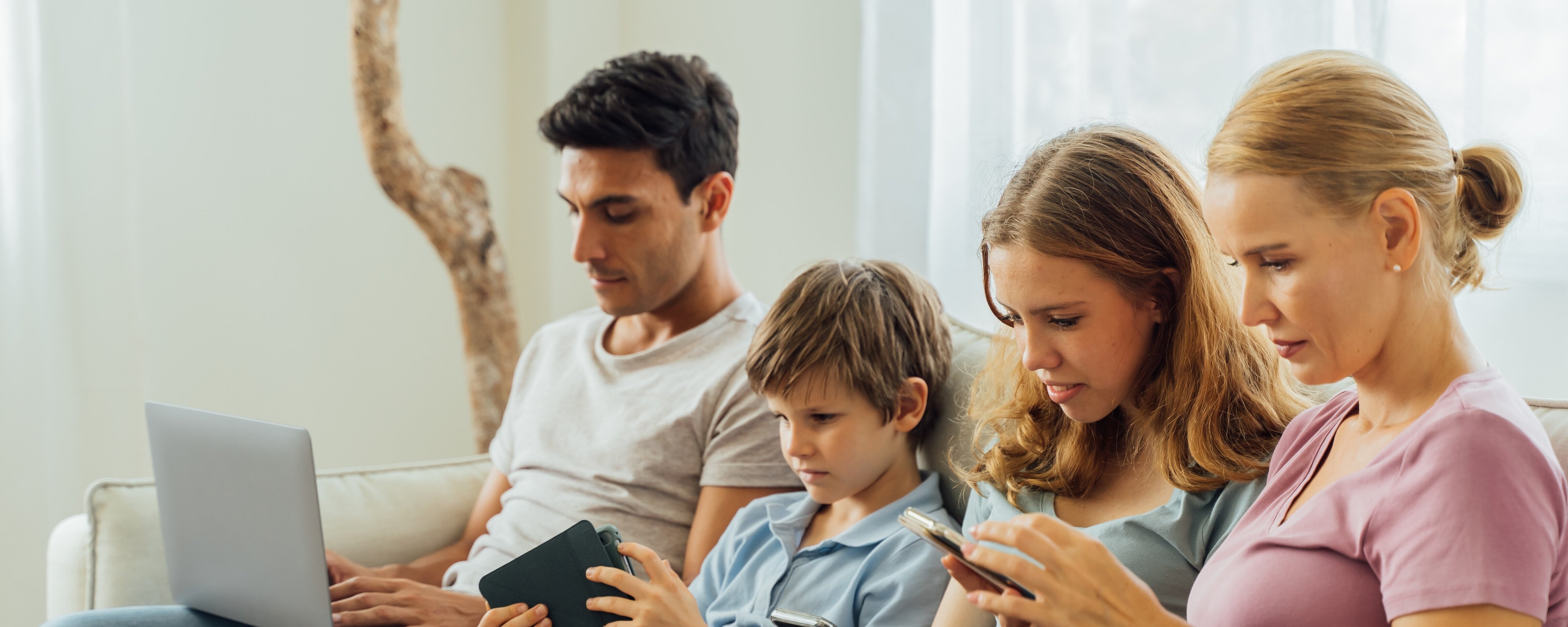 Eine Familie, welche auf einem Sofa sitzt. Jedes Familienmitglied hält ein mobiles Endgerät in den Händen, auf welches es blickt.