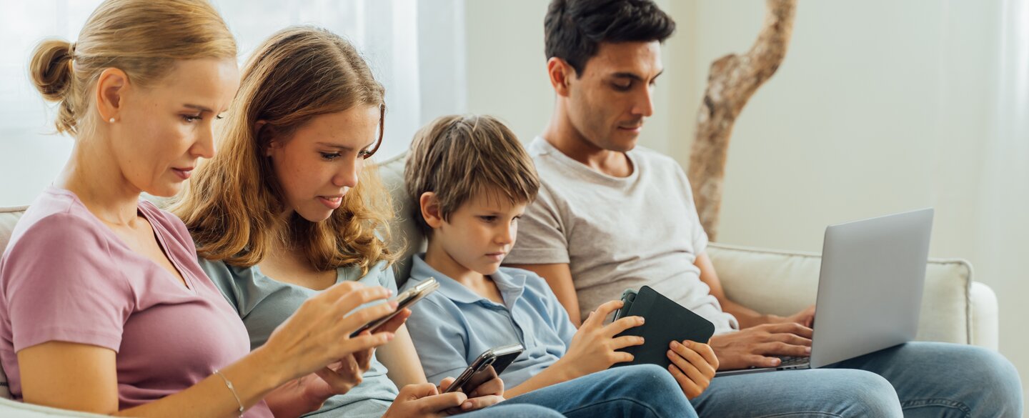 Eine Familie, welche auf einem Sofa sitzt. Jedes Familienmitglied hält ein mobiles Endgerät in den Händen, auf welches es blickt.