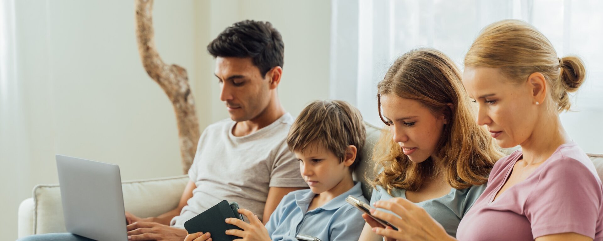 Eine Familie, welche auf einem Sofa sitzt. Jedes Familienmitglied hält ein mobiles Endgerät in den Händen, auf welches es blickt.