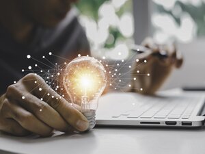 Eine Hand hält eine Glühbirne. Im Hintergrund ist ein Laptop zu sehen.