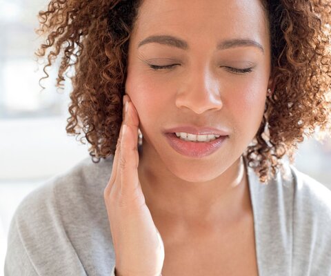 Frau greift sich an die Wange, während Sie ihr Gesicht leicht schmerzhaft verzieht.