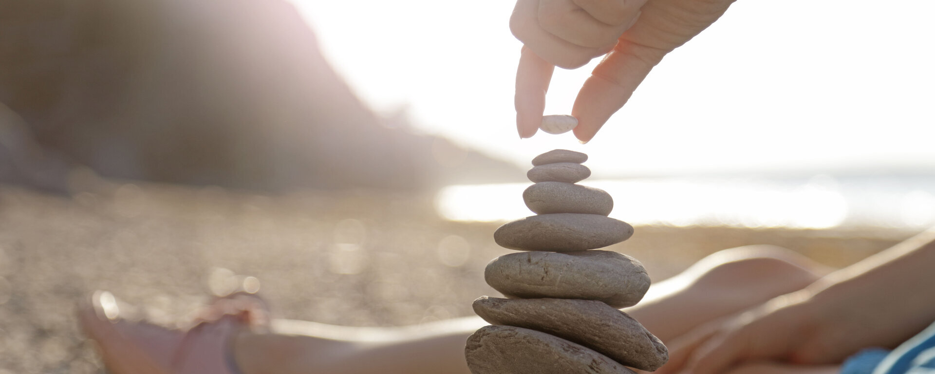 Eine Frau liegt am Strand. Es sind ihre Beine zu sehen und sie stapelt Steine übereinander.