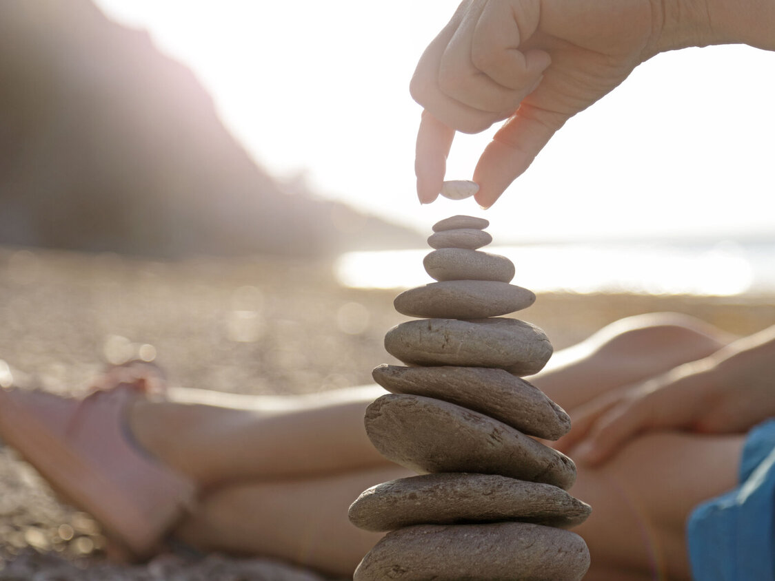 Eine Frau liegt am Strand. Es sind ihre Beine zu sehen und sie stapelt Steine übereinander.