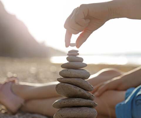 Eine Frau liegt am Strand. Es sind ihre Beine zu sehen und sie stapelt Steine übereinander.