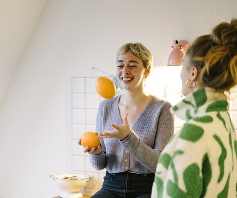 Zwei Frauen stehen in einer Küche und die eine jongliert mit zwei Orangen