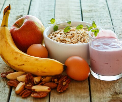 Auf einem Holztisch liegt eine Banane, ein Apfel, Nüsse, Mandarinen, eine Schüssel mit Müsli und ein Glas mit Joghurt.
