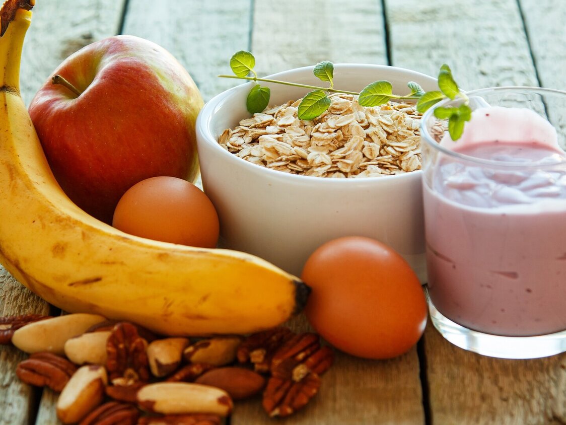 Auf einem Holztisch liegt eine Banane, ein Apfel, Nüsse, Mandarinen, eine Schüssel mit Müsli und ein Glas mit Joghurt.