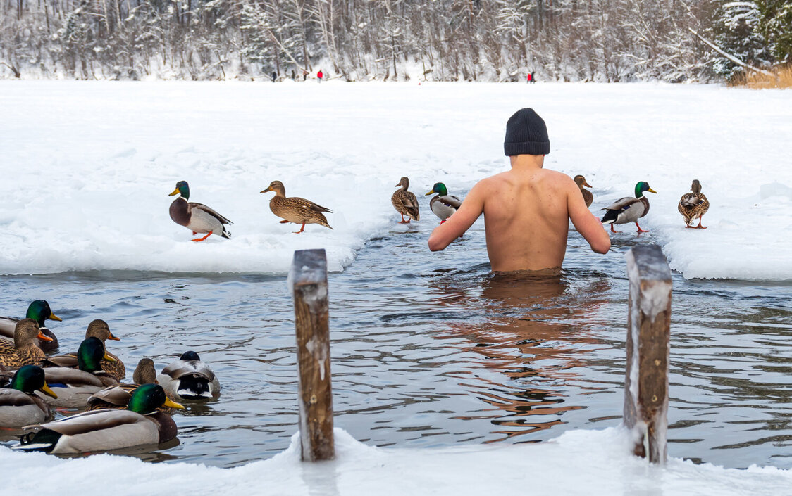 Ein Mann watet ins Wasser, welches von Schnee umgeben ist. Enten befinden sich auf dem Wasser sowie im Hintergrund.