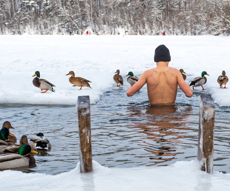 Ein Mann watet ins Wasser, welches von Schnee umgeben ist. Enten befinden sich auf dem Wasser sowie im Hintergrund.