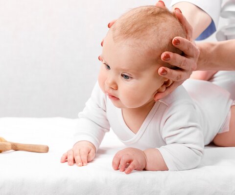 Ein Baby liegt bäuchlings auf einer weißen Unterlage und sein Kopf wird von hinten mit zwei Händen umfasst.