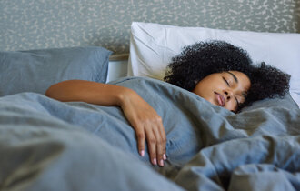 Eine dunkelhäutige Frau liegt unter einer grauen Decke im Bett und schläft.