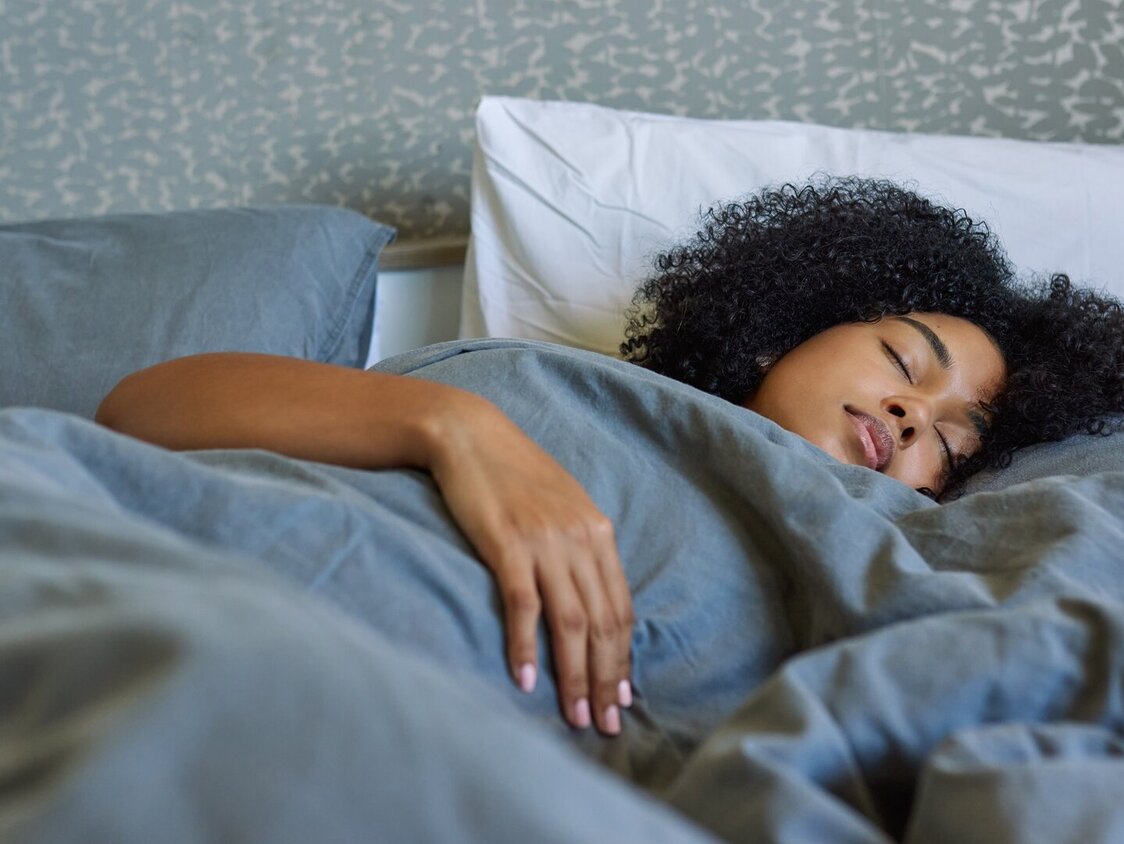 Eine dunkelhäutige Frau liegt unter einer grauen Decke im Bett und schläft.