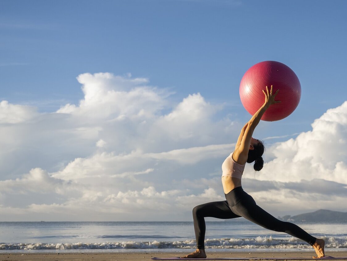 Eine Frau macht am Strand eine Yoga-Übung und hält dabei einen Gymnastikball über den Kopf.