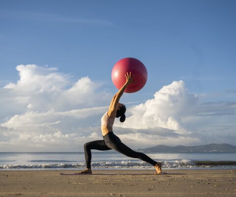 Eine Frau macht am Strand eine Yoga-Übung und hält dabei einen Gymnastikball über den Kopf.