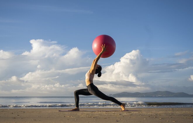 Eine Frau macht am Strand eine Yoga-Übung und hält dabei einen Gymnastikball über den Kopf.