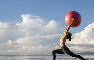 Eine Frau macht am Strand eine Yoga-Übung und hält dabei einen Gymnastikball.