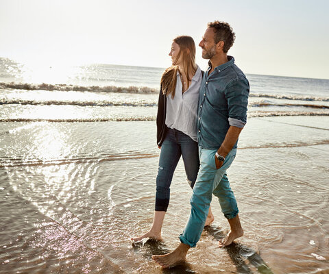  Ein Paar bei einem Spaziergang am Strand. Sie laufen barfuß durch das Wasser. Meer und Sonne im Hintergrund.