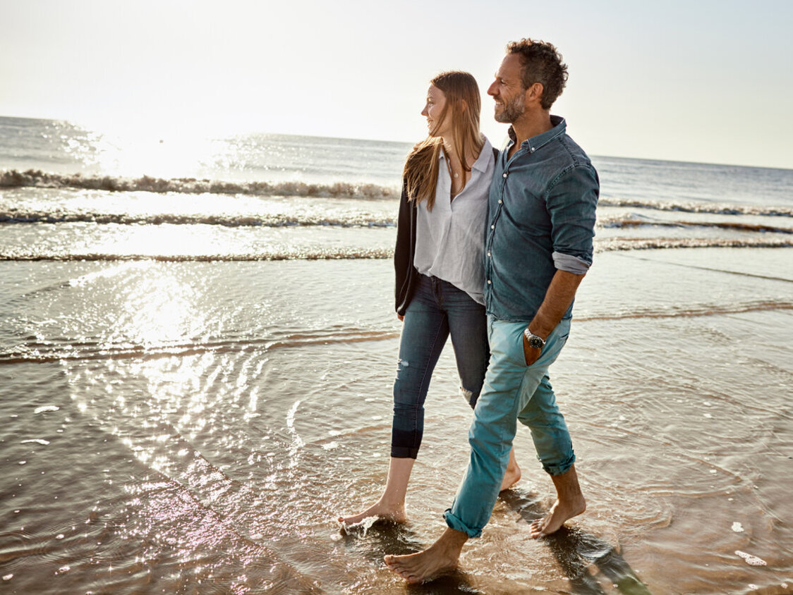  Ein Paar bei einem Spaziergang am Strand. Sie laufen barfuß durch das Wasser. Meer und Sonne im Hintergrund.