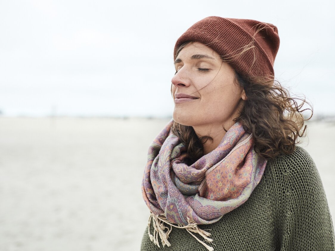 Eine Frau mit Mütze und Schal steht mit geschlossenen Augen am Strand.
