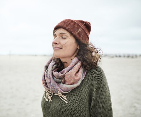 Eine Frau mit Mütze und Schal steht mit geschlossenen Augen am Strand.