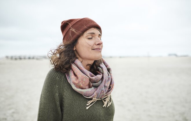 Eine Frau mit Mütze und Schal steht mit geschlossenen Augen am Strand.