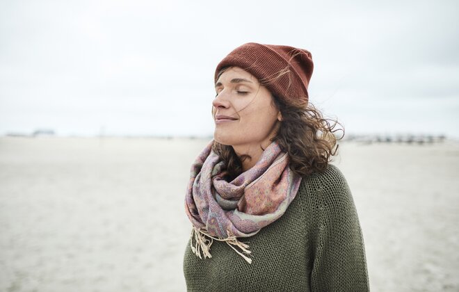 Eine Frau mit Mütze und Schal steht mit geschlossenen Augen am Strand.