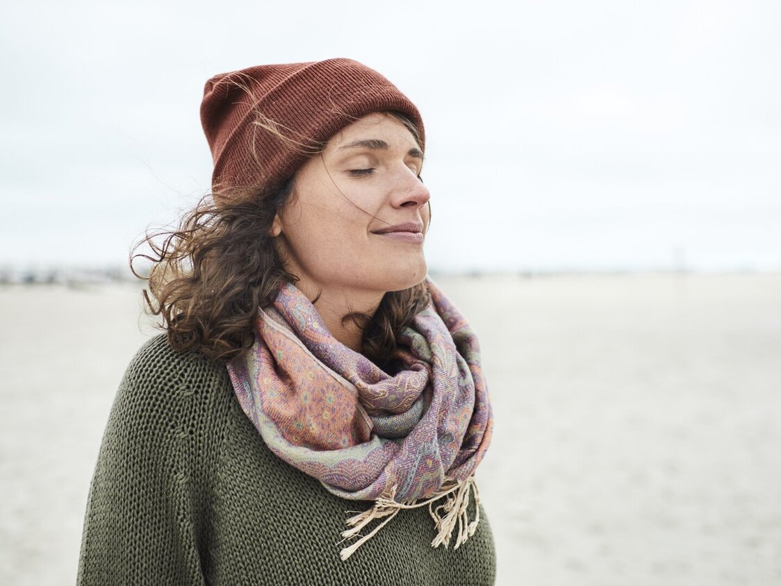 Eine Frau mit Mütze und Schal steht mit geschlossenen Augen am Strand.