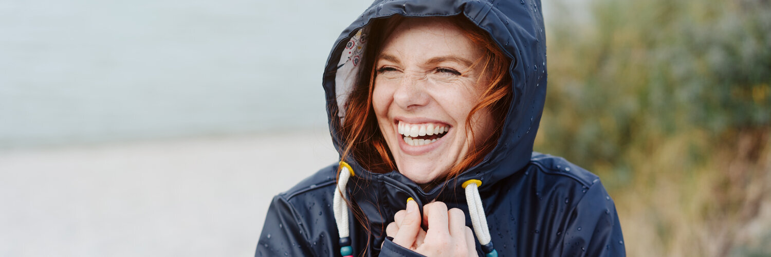 Eine Frau in einer Jacke mit aufgezogener Kapuze. Sie hält die Jacke fest und lacht. Im Hintergrund Strand.