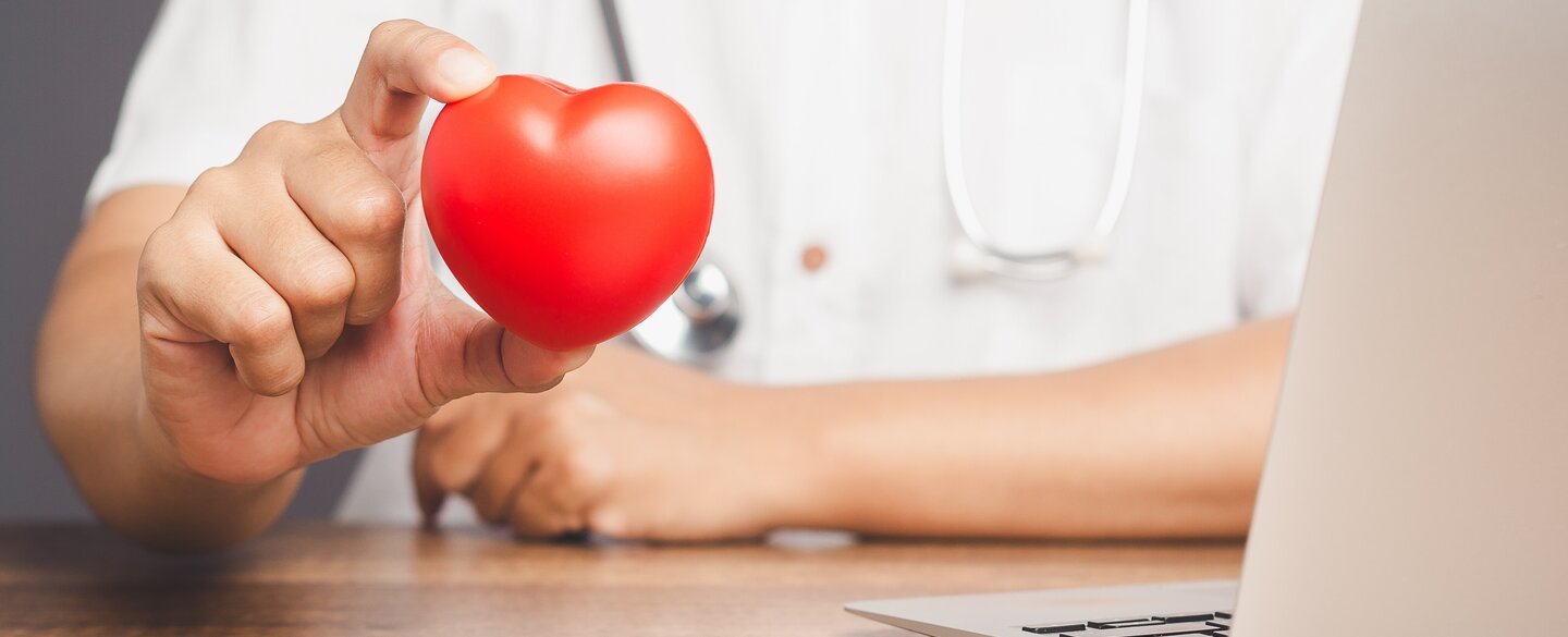 Ein Arzt sitzt vor einem Laptop und hält ein Herz hoch.