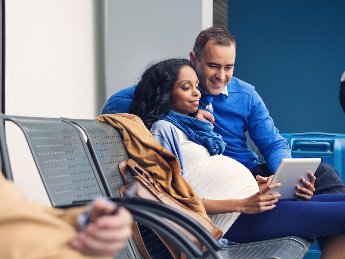 Eine Schwangere Frau sitzt neben einem Mann an einem Flughafen und zeigt ihm etwas auf dem Tablet.